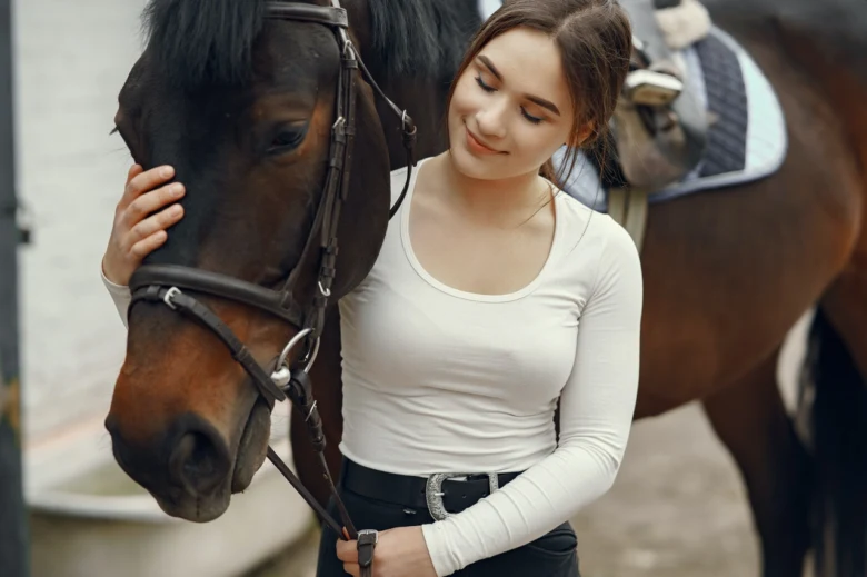 Žena hladí hnědého koně u stájových pomůcek.